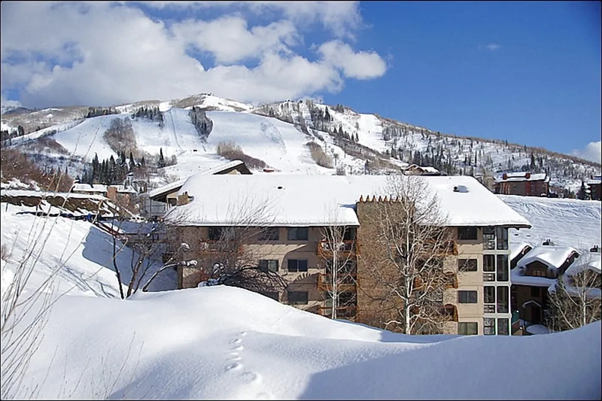 A wide view of snow-covered slopes with a mountain backdrop and the building’s architecture in the foreground. Tracks in the snow suggest recent skiing and snowboarding activity, while the crisp morning light casts a serene glow over the scene.