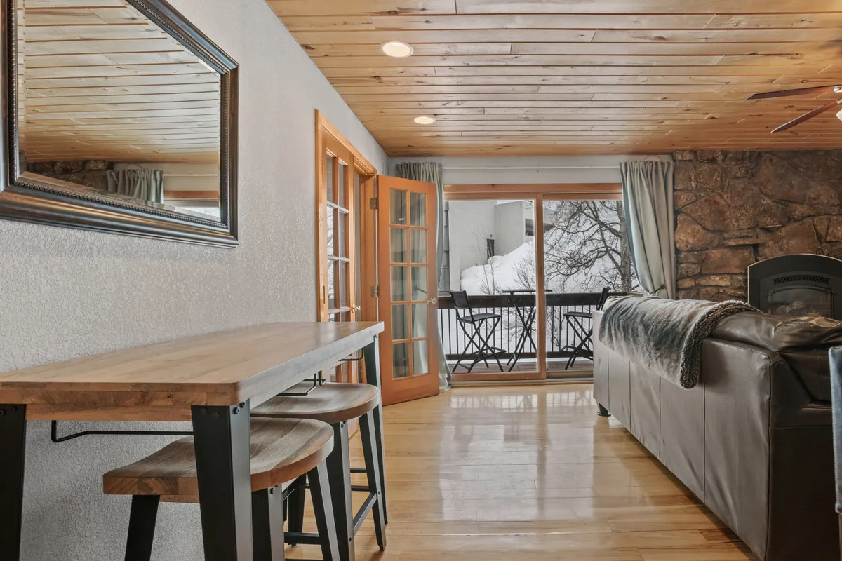 A cozy dining area featuring a sturdy wooden table set near a sliding glass door, with a stone fireplace and leather couch in view. The arrangement suggests a warm and inviting space for meals, relaxation, and enjoying the surrounding scenery.
