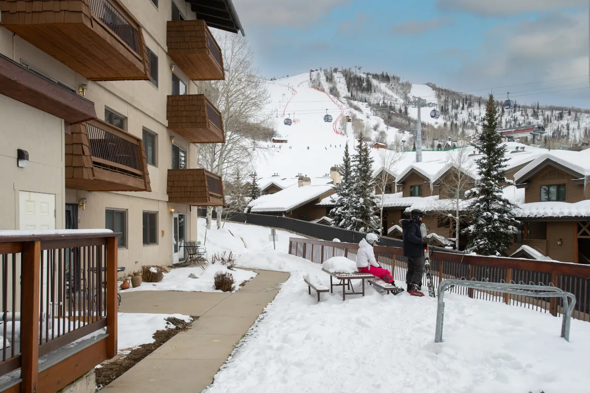 A cheerful winter scene with the condo’s balconies overlooking a bustling ski slope, where families and skiers prepare for a day outdoors. The snowy landscape and visible building emphasize ease of access and the vibrant atmosphere at the base area.