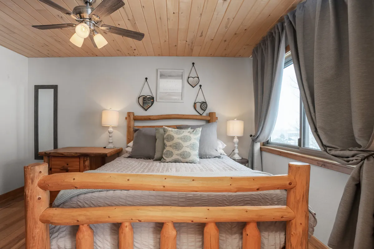 A tranquil bedroom featuring a log bed with fine linens, decorative pillows, nightstands with lamps, and a window with curtains allowing in natural light. Decorative wall art enhances the serene and inviting atmosphere of the space.
