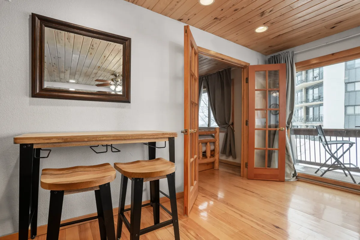A relaxed interior view featuring a wooden bar table and stools, with open doors leading directly to a private balcony. The setup invites guests to step outside, blending interior comfort with outdoor access.