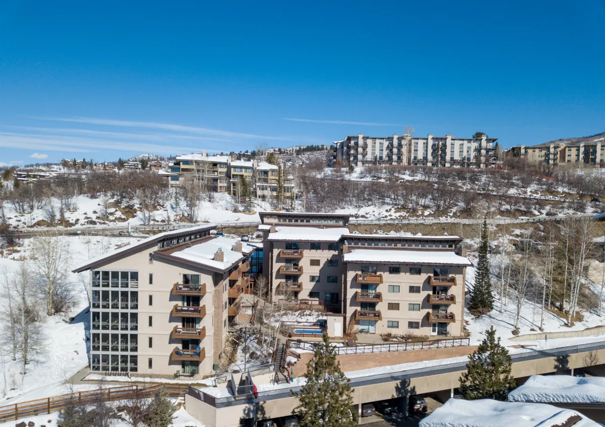 A view of the building’s modern architecture is set within a snowy landscape and tall trees. The scene suggests access to winter sports and a cozy retreat after a day outdoors.