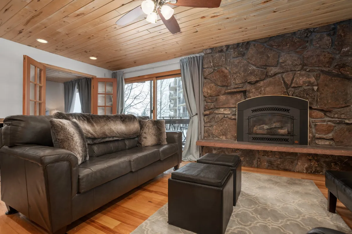 A warm and inviting living room featuring a plush sofa positioned near a striking stone fireplace, set against large windows that let in natural light. The hardwood floors and comfortable furnishings make it an ideal space to relax, read, or gather for a movie night.