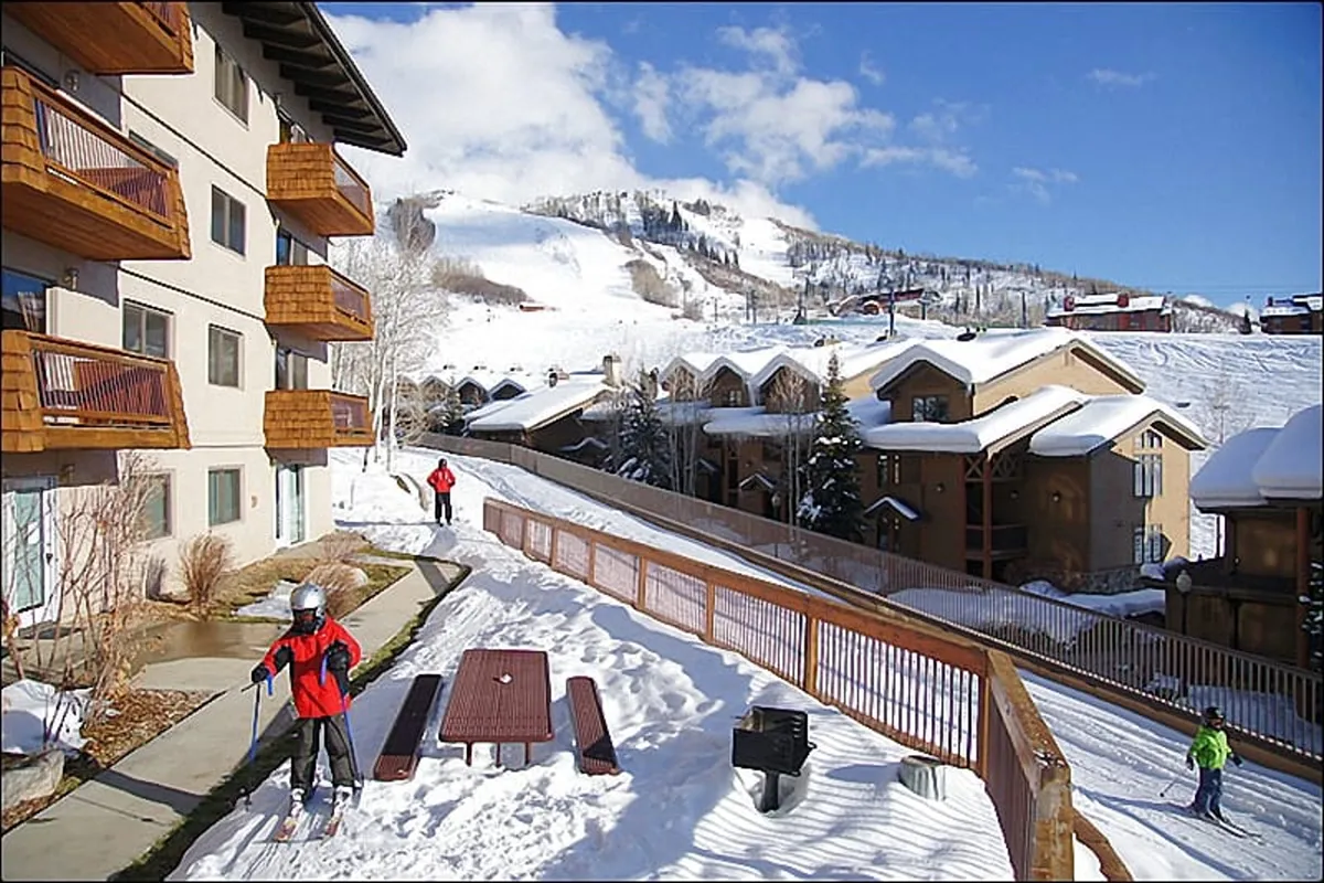 The building’s balconies overlook a playful winter scene where skiers traverse heated sidewalks, with a backdrop of snow-covered mountains. A picnic table hints at social gatherings and year-round outdoor enjoyment.