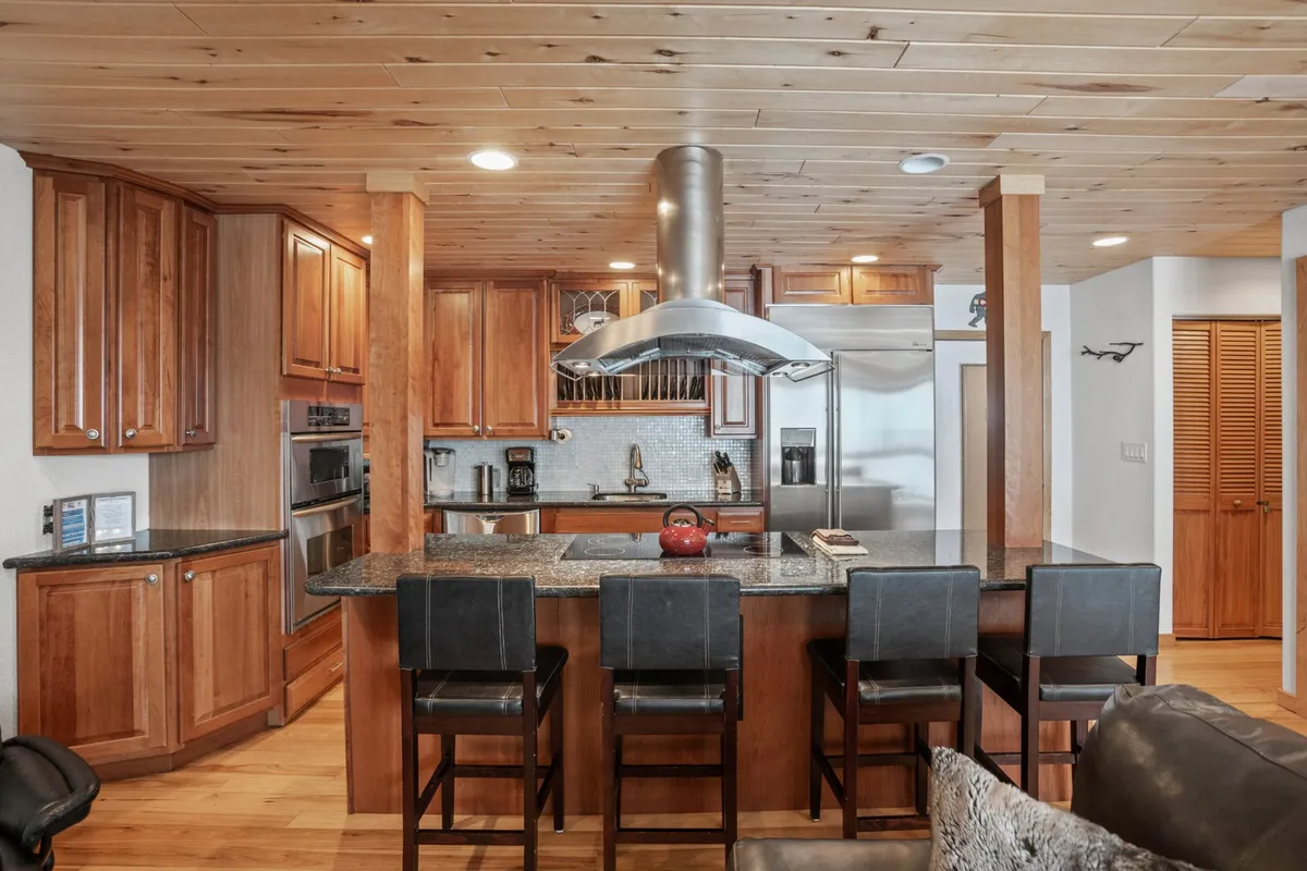 This image features a central kitchen island surrounded by stainless steel appliances, warm wood cabinetry, and a sleek modern range hood. The space is arranged to facilitate both cooking and gathering, with ample counter space and inviting lighting that creates a cozy, welcoming atmosphere.