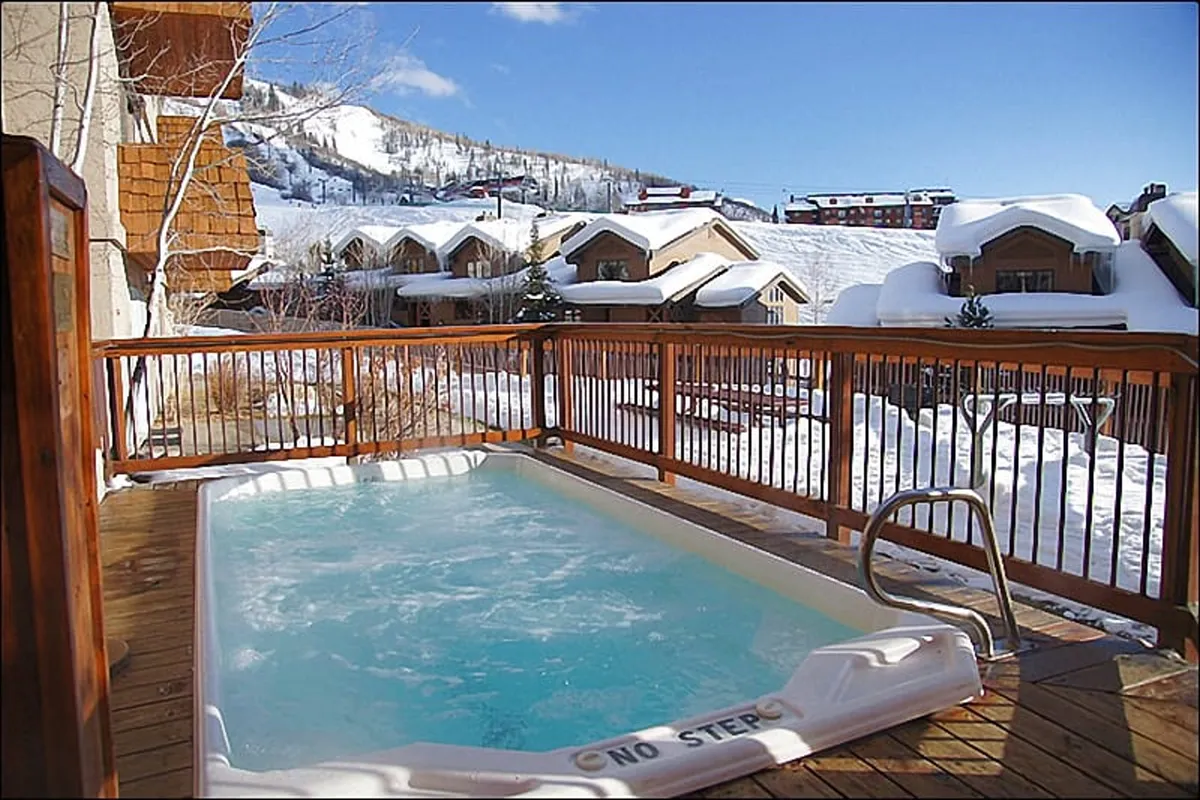 An inviting outdoor hot tub sits on a wooden deck surrounded by snow-covered mountains and cozy cabins. The scene evokes relaxation and a sense of post-adventure comfort, with the hot tub appearing ready for use after a day on the slopes.