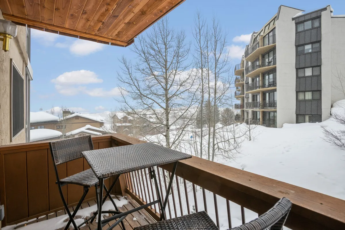 A cozy balcony with outdoor seating, looking out onto a snowy landscape and adjacent buildings. The scene evokes a sense of peacefulness and comfort, with fresh snow hinting at recent mountain adventures and a perfect spot for a warm drink or morning yoga.