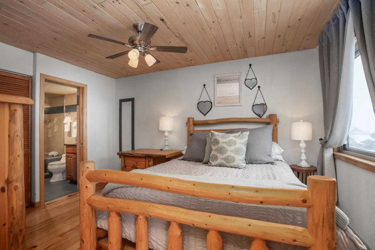 A calm and comfortable bedroom with a wooden bed frame, decorative pillows, nightstands with lamps, framed wall art, and a window with curtains. The space is bathed in soft, natural light, evoking a sense of peaceful retreat.