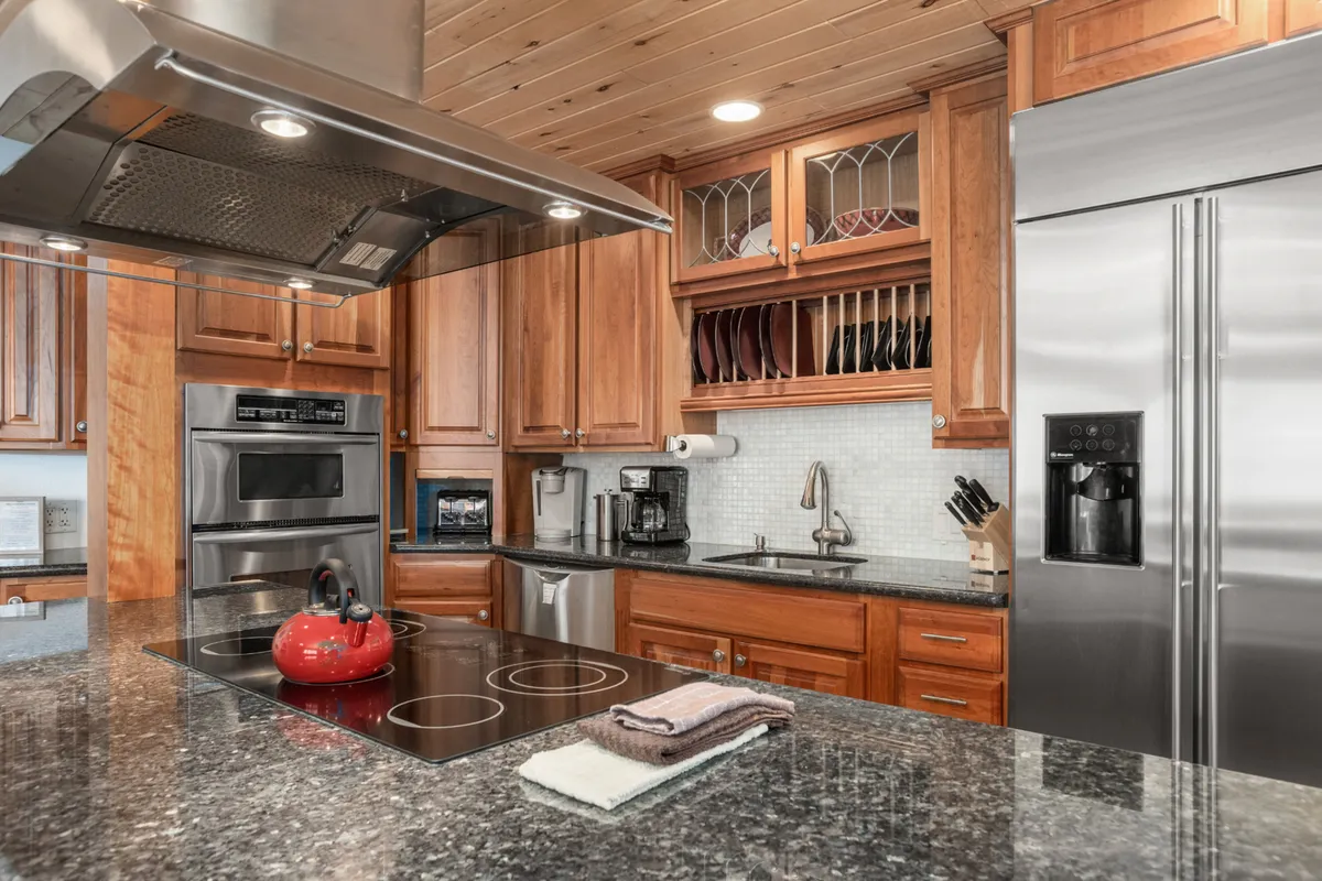 This inviting kitchen features a spacious island topped with a bright red kettle, complemented by sleek stainless steel appliances and rich wood cabinetry with glass-fronted accents. The warm, welcoming design encourages shared meals, morning coffee rituals, and quality time spent after a day on the Steamboat Springs slopes.