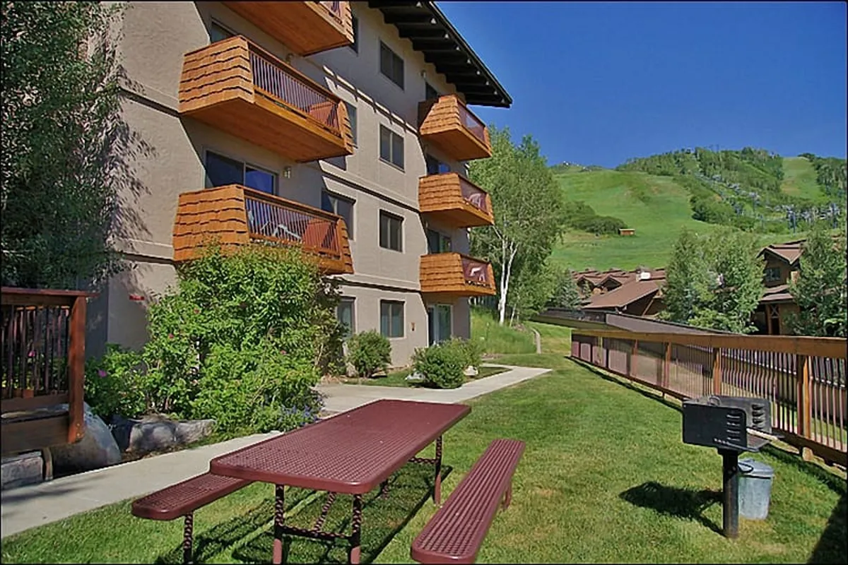 A sunny exterior view featuring balconies, a picnic table set on a green lawn, and a panoramic mountain backdrop. The inviting outdoor space suggests summer activities like picnics, relaxing, and easy trail access for hiking or biking right outside the door.